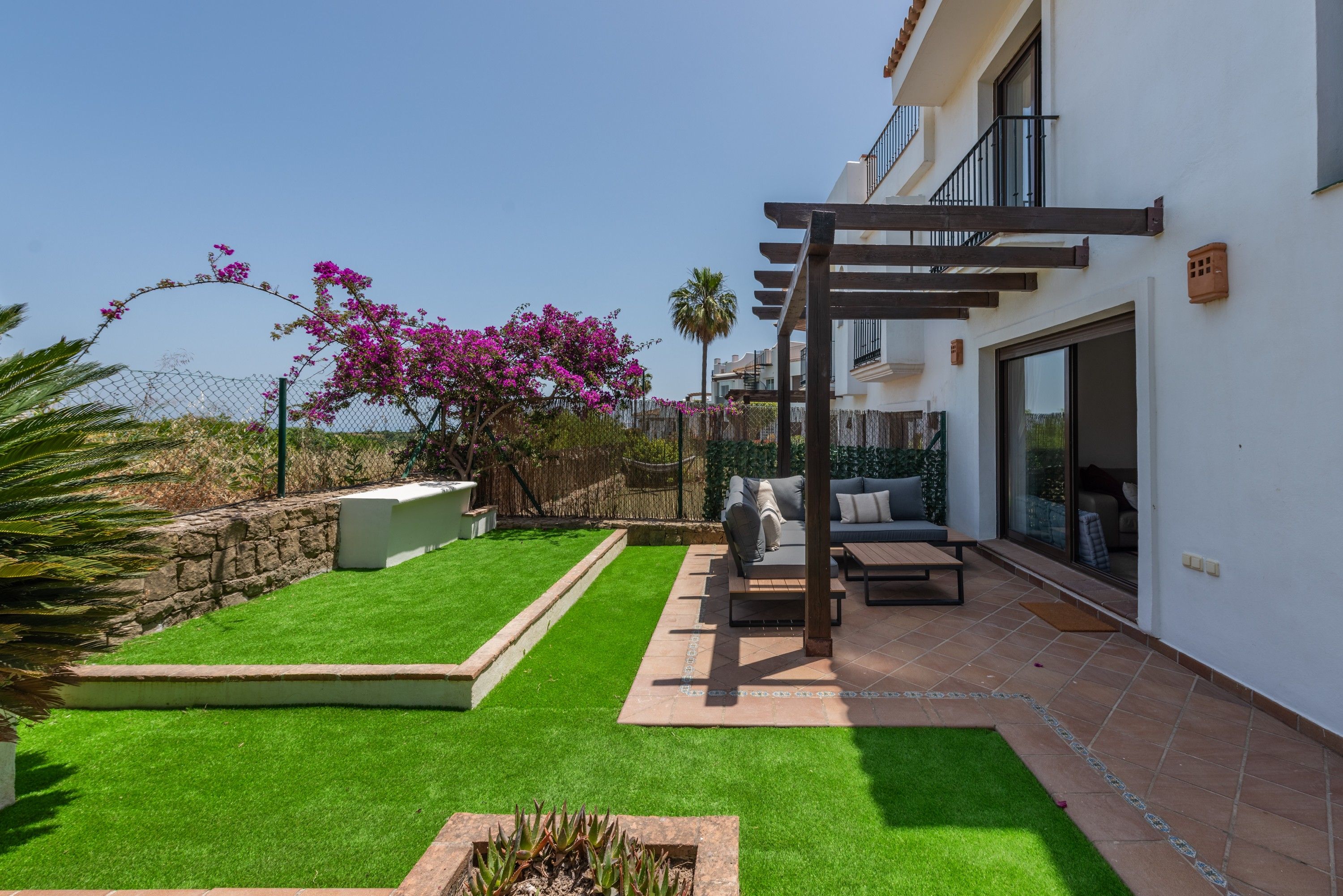 Une terrasse résidentielle avec une pelouse artificielle, une pergola, du mobilier d’extérieur et un mur en pierre.