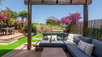 Un espace extérieur aménagé, abrité et doté d’un canapé et d’une table, entouré d’un jardin avec des plantes en fleurs et un mur en pierre.
