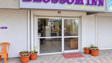 The exterior of a hotel named 'Blossom Inn' with a sign indicating 'Safe & Comfortable Stay.' There are three potted plants in front of the entrance, and a 'Welcome' mat is visible on the ground.