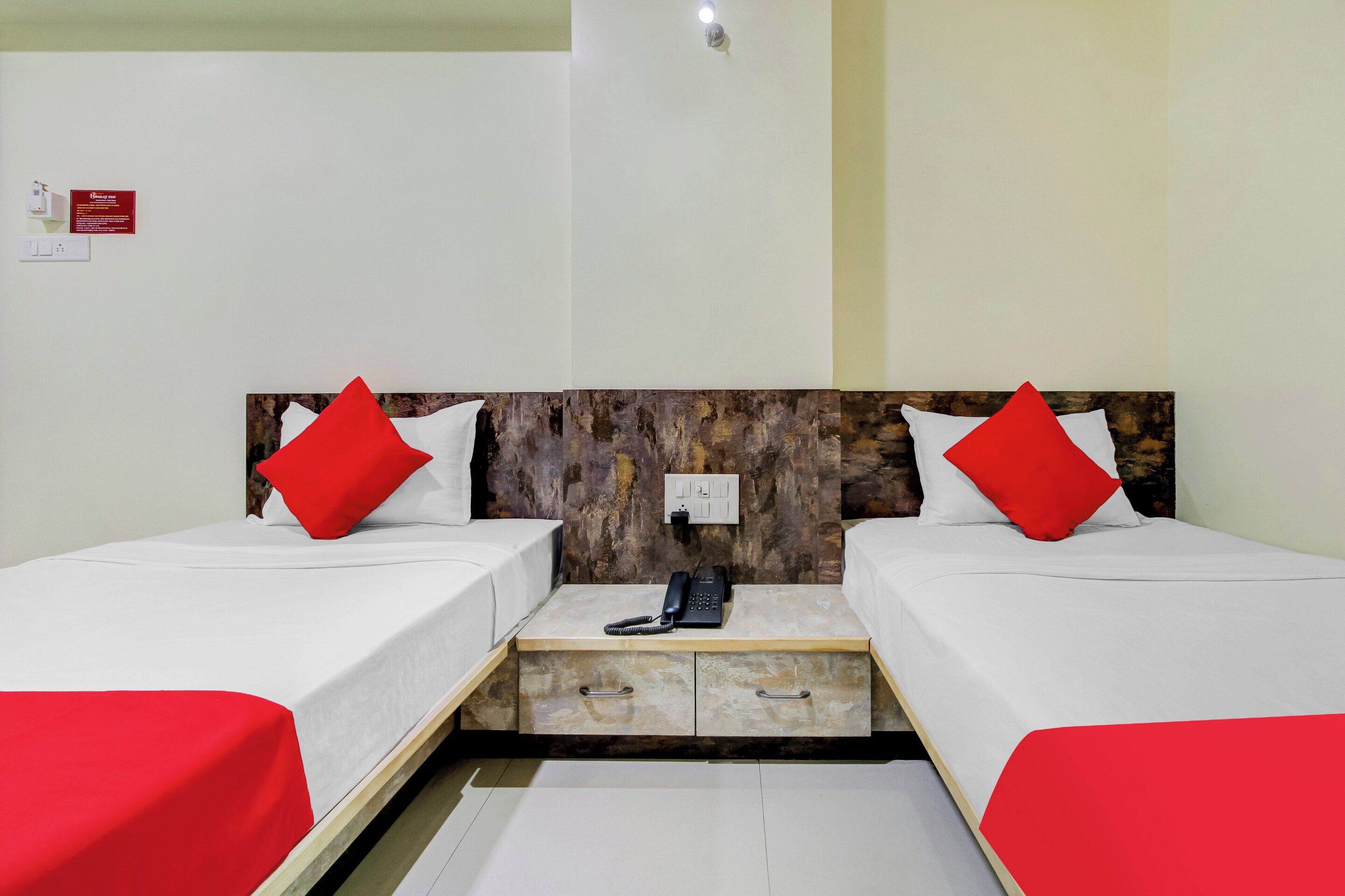 Une chambre d’hôtel avec deux lits, chacun agrémenté d’un oreiller rouge, une table de chevet avec un téléphone et un luminaire fixé au mur.