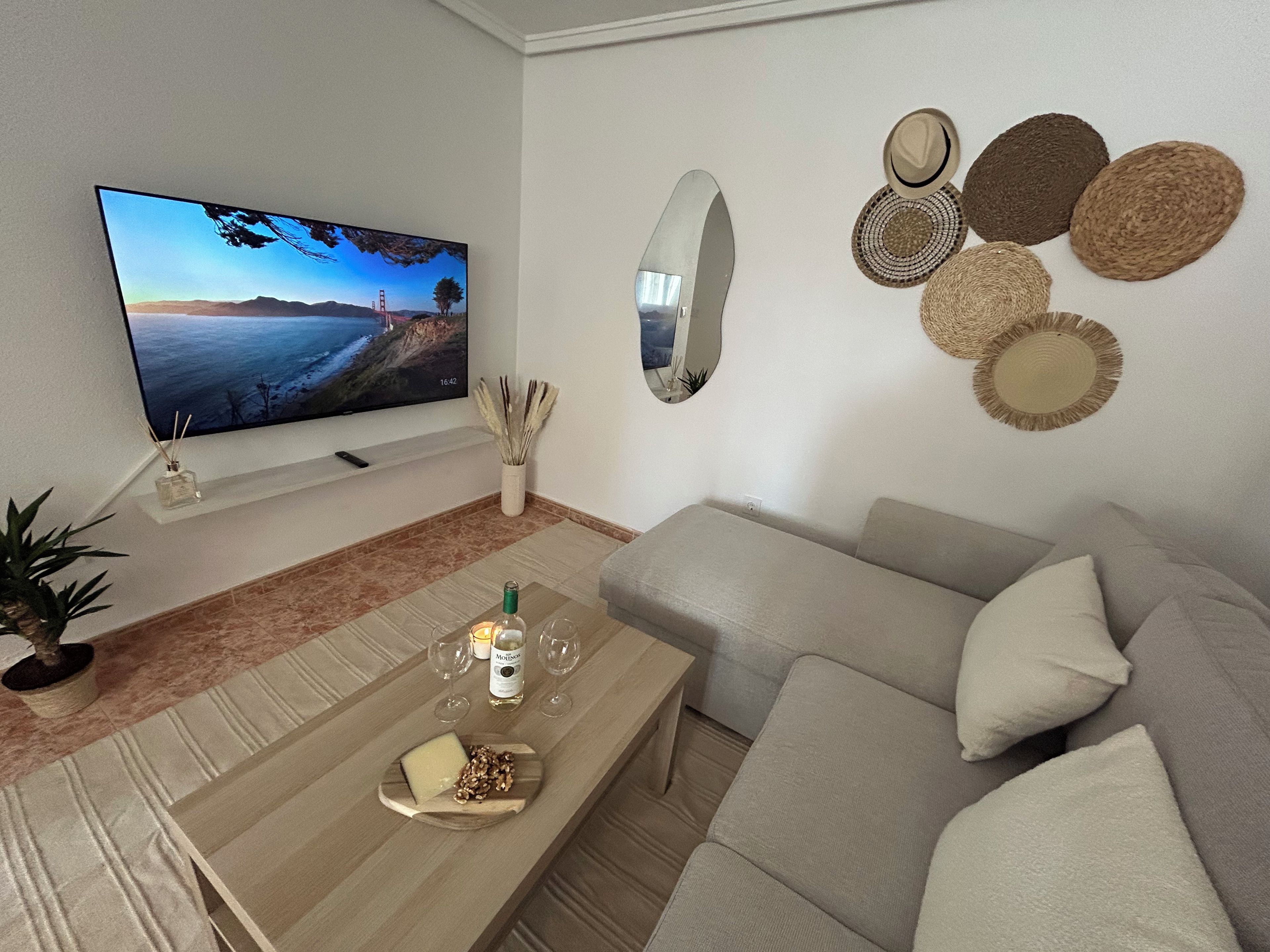 A living room with a flat-screen TV, a sofa, a coffee table with snacks, a mirror, and decorative wall hangings.