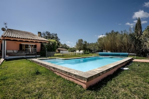 A rectangular swimming pool with a stone edge, surrounded by a grassy lawn, a small building with a tiled roof, and trees in the background. - Villa 'Unique" With Pool - Sea & Mountain View' (Port de Pollença)