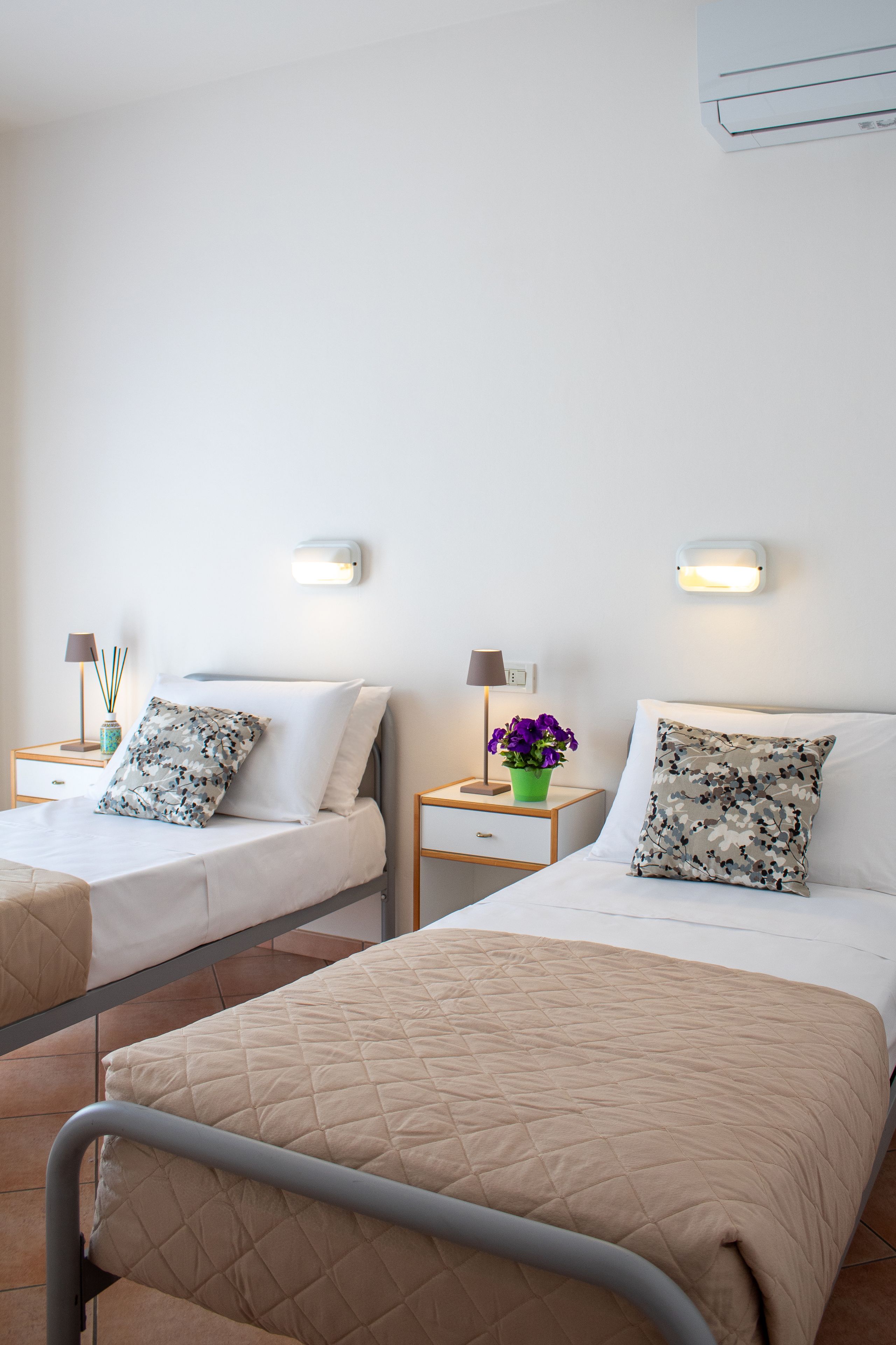 A hotel room with two beds, each with a pillow and a nightstand with a lamp and a vase of flowers.