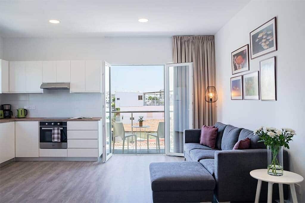 A modern apartment with a kitchenette, a grey sofa, a small table with a vase, and a balcony with a view.