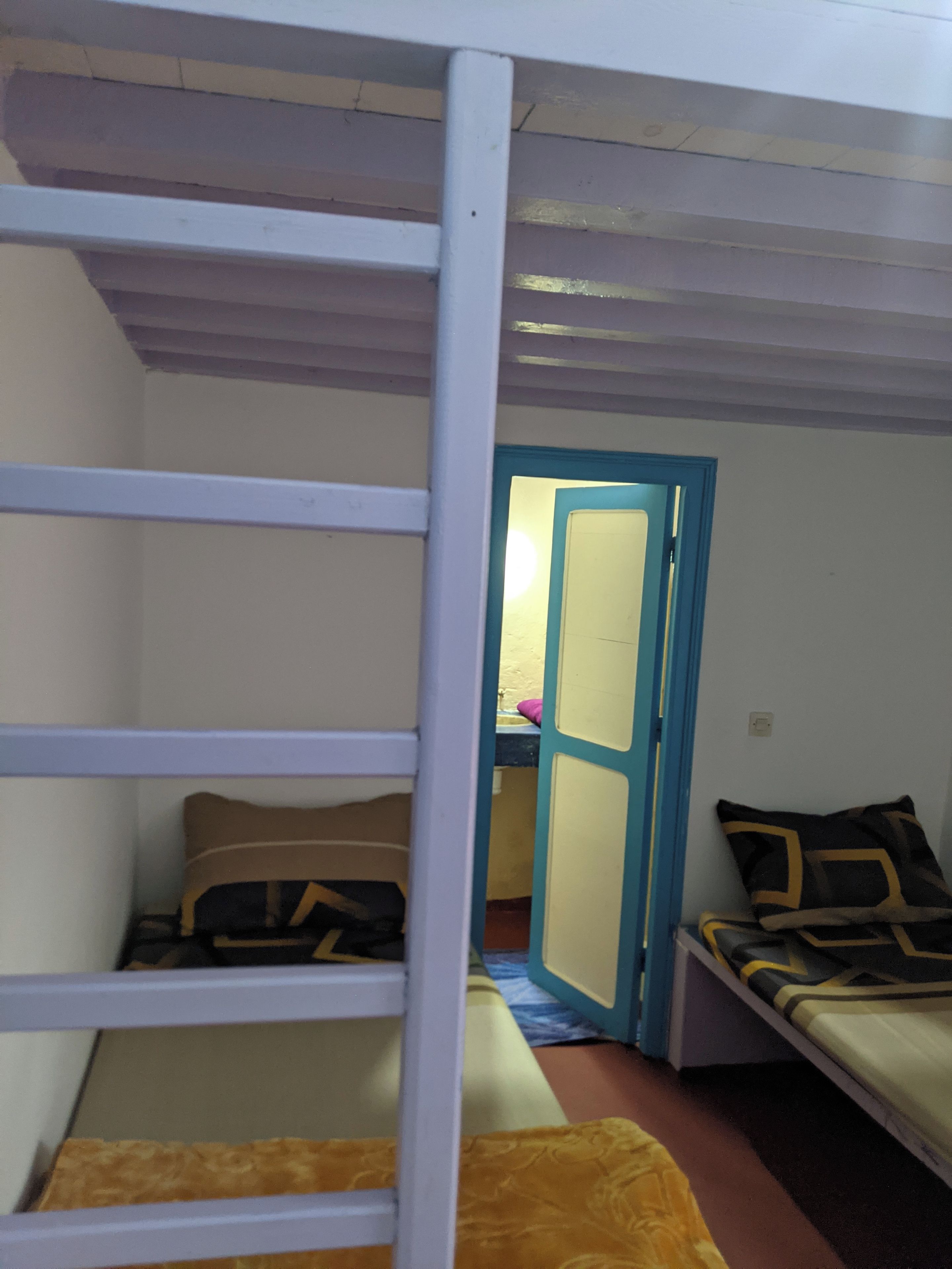Una cama litera con escalera, una mesita con una silla, y una puerta que da a otra habitación.