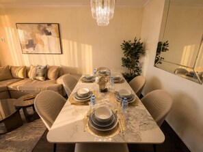 A dining area with a long table set for four, surrounded by chairs, a sofa, and a plant. - Heart of Pearl 3BR, Huge Terrace & Marina Views (Lusail)