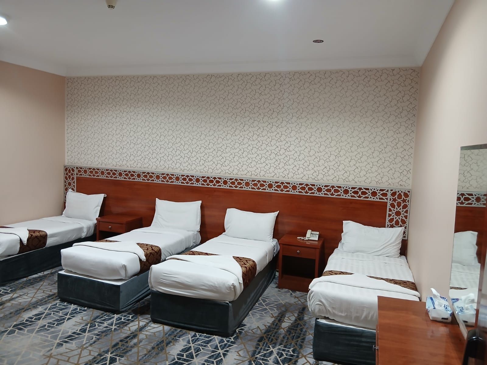 A hotel room with three single beds, a wooden headboard, a patterned carpet, and a small bedside table with a phone.