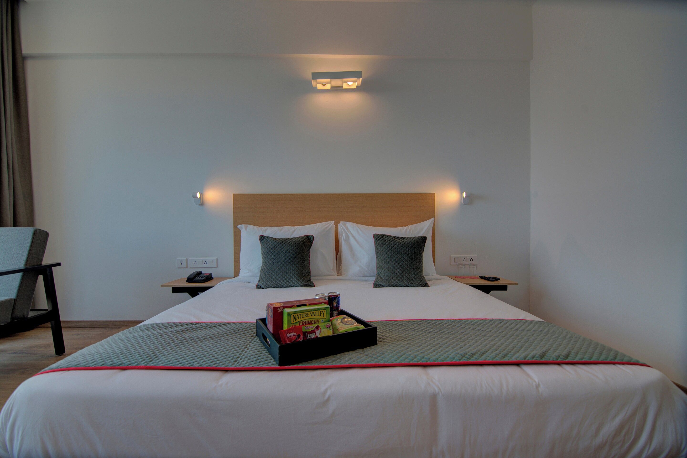 A hotel room with a bed, bedside tables, a chair, and a tray with snacks.