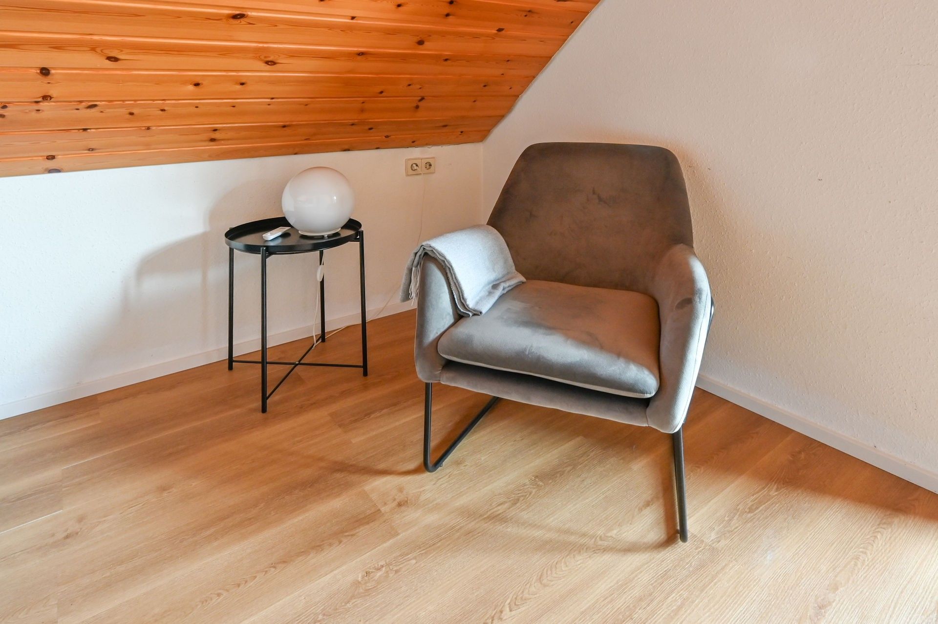 Une pièce moderne avec un plafond en pente, un fauteuil à revêtement gris, une petite table d’appoint noire sur laquelle repose un objet sphérique blanc, et un plancher en bois.