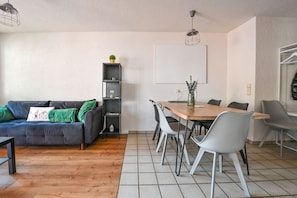 Ein modernes Wohnzimmer mit einem dunkelgrauen Sofa, einem Holzespeisetisch mit Stühlen, einem hohen schwarzen Bücherregal und einer Pendelleuchte.
