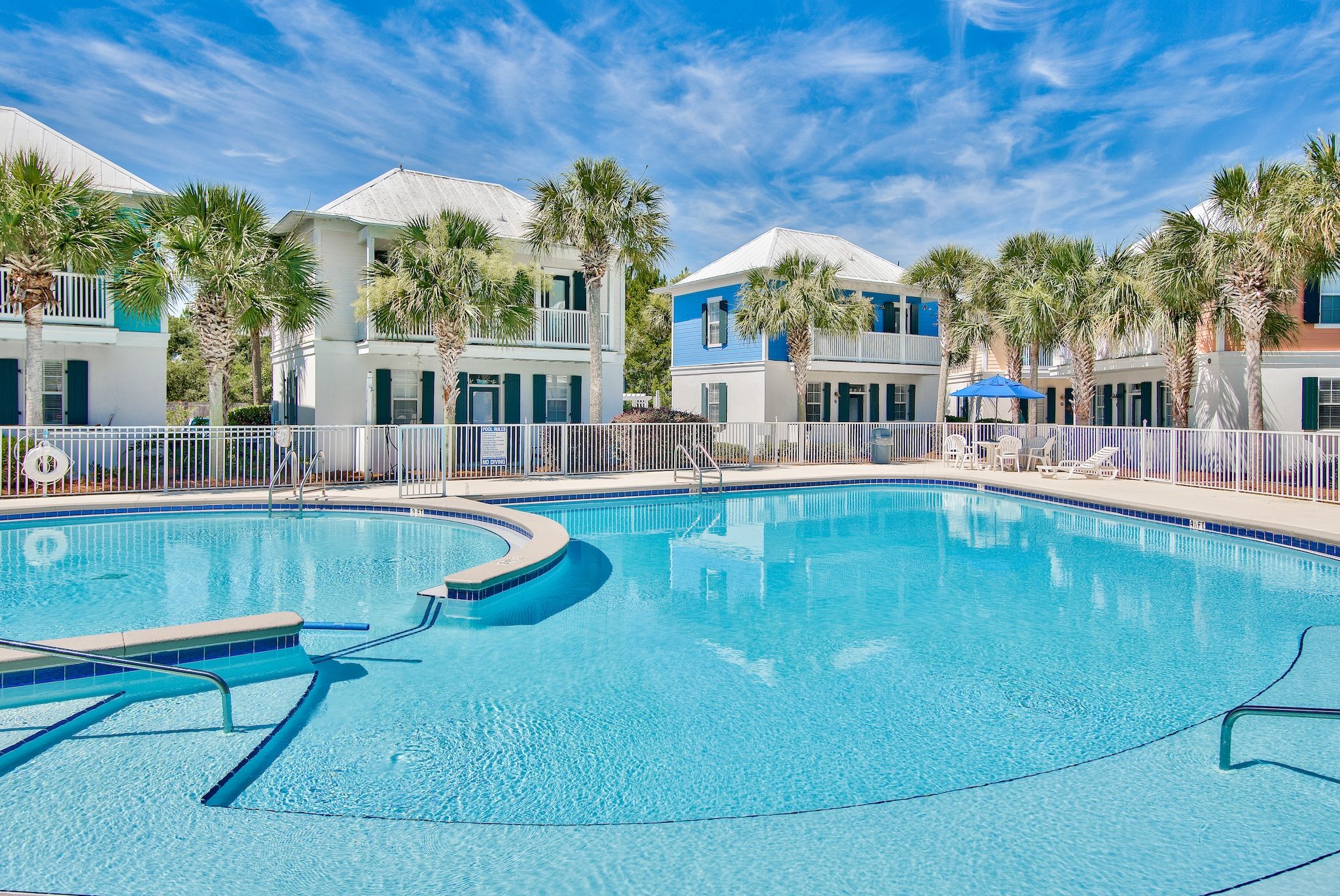 A swimming pool with a white fence, palm trees, and buildings with balconies.