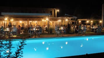 An outdoor dining area with tables and chairs, a pool, and a building with illuminated windows in the background.