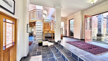 A spacious interior with a staircase leading to a wooden balcony, stone flooring, and a view of the outside.