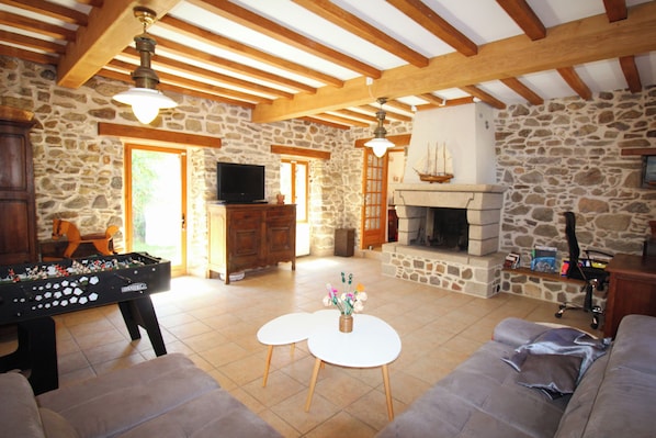A cozy living room with a stone wall, wooden beams, a fireplace, a television, and a foosball table.