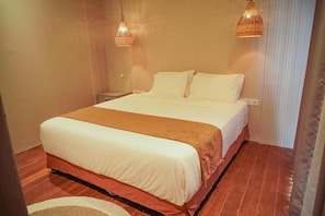 A neatly made bed with a white and brown bedspread, a wooden nightstand with a lamp, and a window with blinds. - Hu'u Beach Front Hotel (Nusa Tenggara Barat)