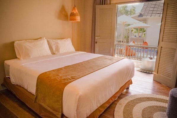 A bedroom with a large bed, a wooden floor, and a view of an outdoor area with umbrellas. - Hu'u Beach Front Hotel (Nusa Tenggara Barat)
