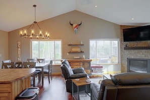 A spacious living room with a fireplace, a dining area with a chandelier, and a large window offering a view of the outdoors.
