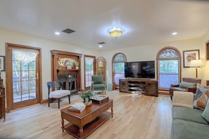A spacious living room with a fireplace, comfortable seating, a flat-screen TV, and large windows.