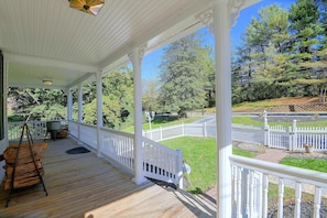 A covered porch with a wooden deck, white railings, and a view of a landscaped garden with trees and a fence.