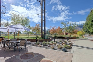Un patio al aire libre con mesas y sillas, un sendero de piedra y vistas a un estacionamiento y edificios al fondo.