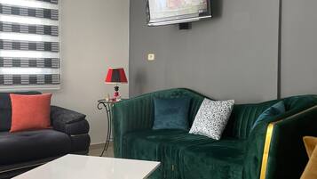 Un soggiorno moderno con un divano verde, un divano nero, un tavolino da caffè bianco, una poltrona gialla, una TV a parete e una finestra con persiane.