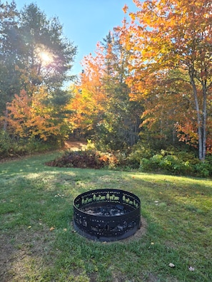 A circular fire pit with a metal rim and a central fire, surrounded by grass and trees with autumn foliage. - Enjoy a private home on 5 acres, abutting Jordan River. Keyless entry and Wi-Fi. (Lamoine)