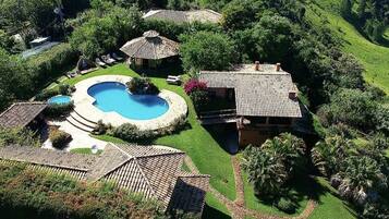 A large swimming pool surrounded by a well-maintained lawn, with multiple buildings and a hilly landscape in the background.