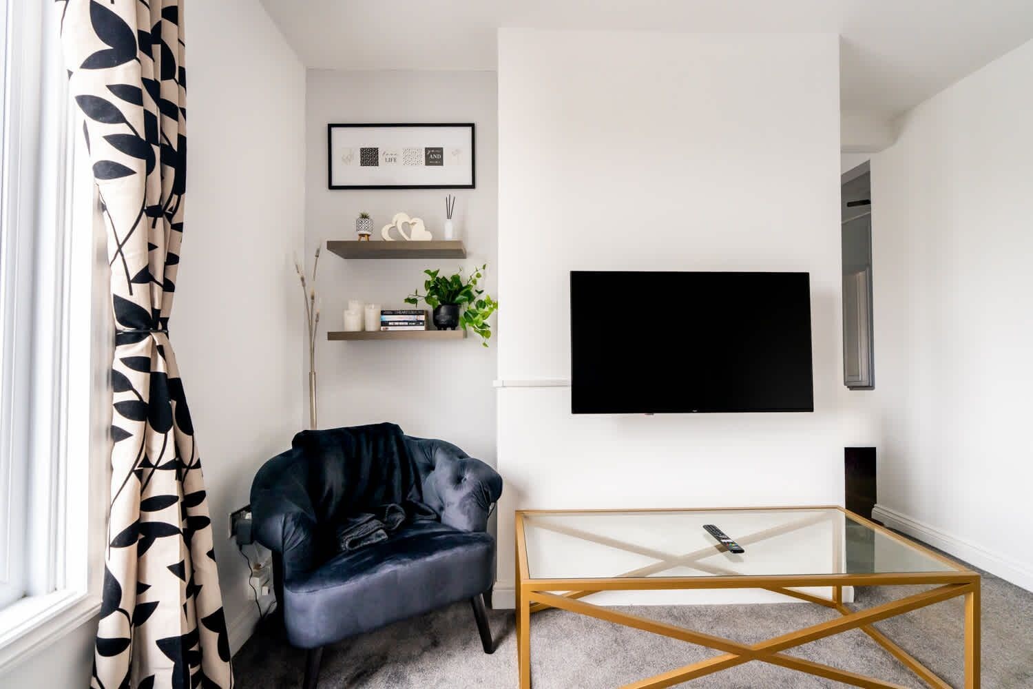 Ett vardagsrum med en svart lädersoffa, ett soffbord i glas, en platt-TV som är monterad på väggen och ett fönster med gardiner.