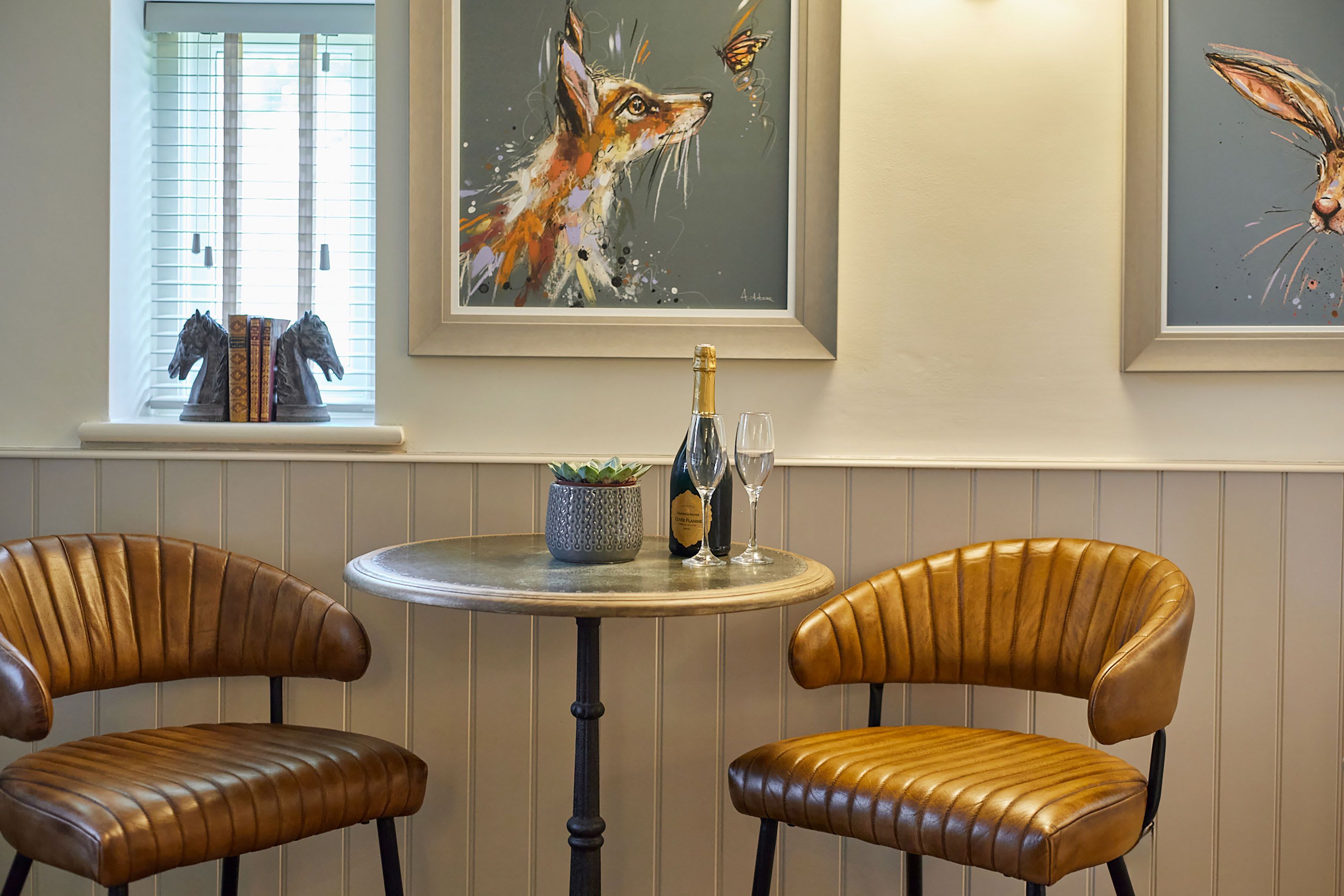 A room with two leather chairs, a round table with a bottle and glasses, and three framed artworks on the wall.