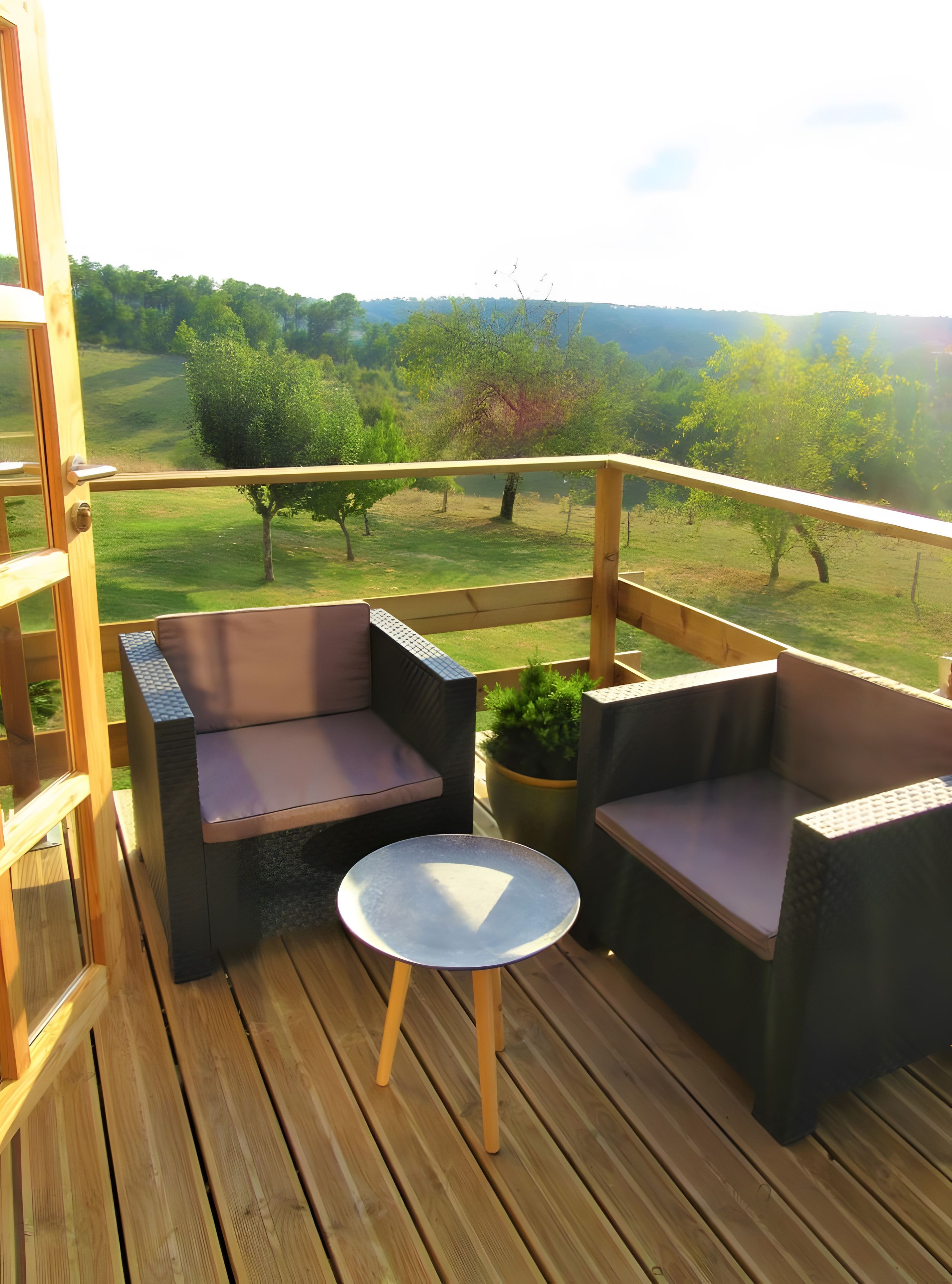 A wooden deck with two cushioned seating areas and a small table, overlooking a grassy landscape with trees.