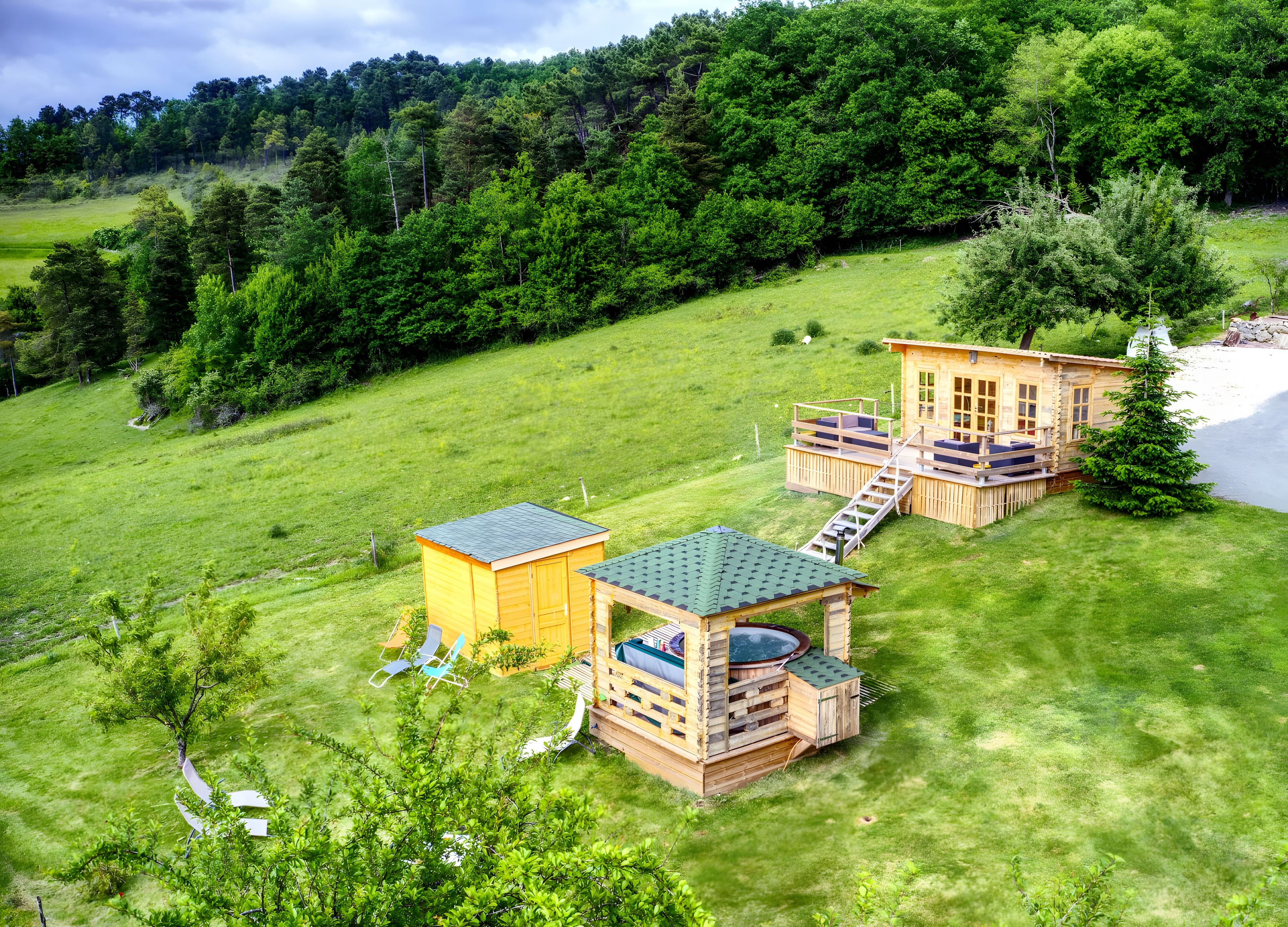 A wooden cabin with a deck, a small shed, and a hot tub in a grassy field with a forested hill in the background.