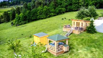A wooden cabin with a deck, a small shed, and a hot tub in a grassy field with a forested hill in the background.