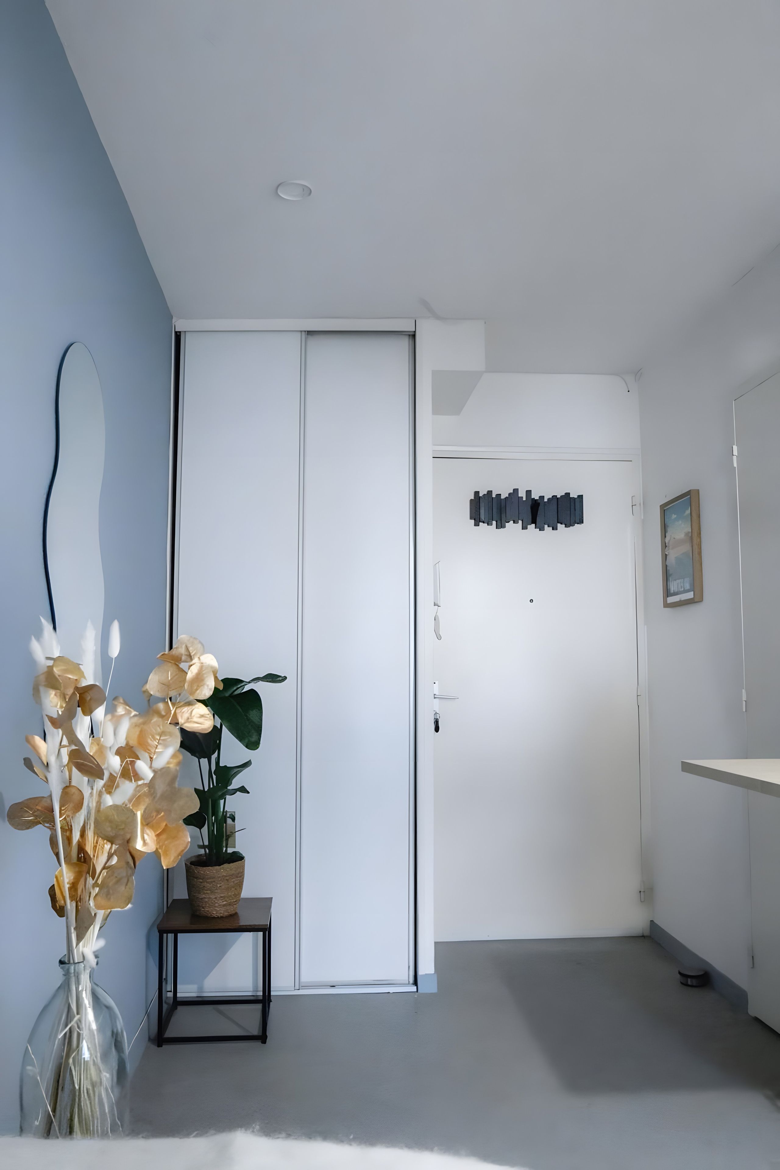 Un couloir avec une porte blanche, une œuvre d’art accrochée au mur et une plante en pot posée sur une petite table.