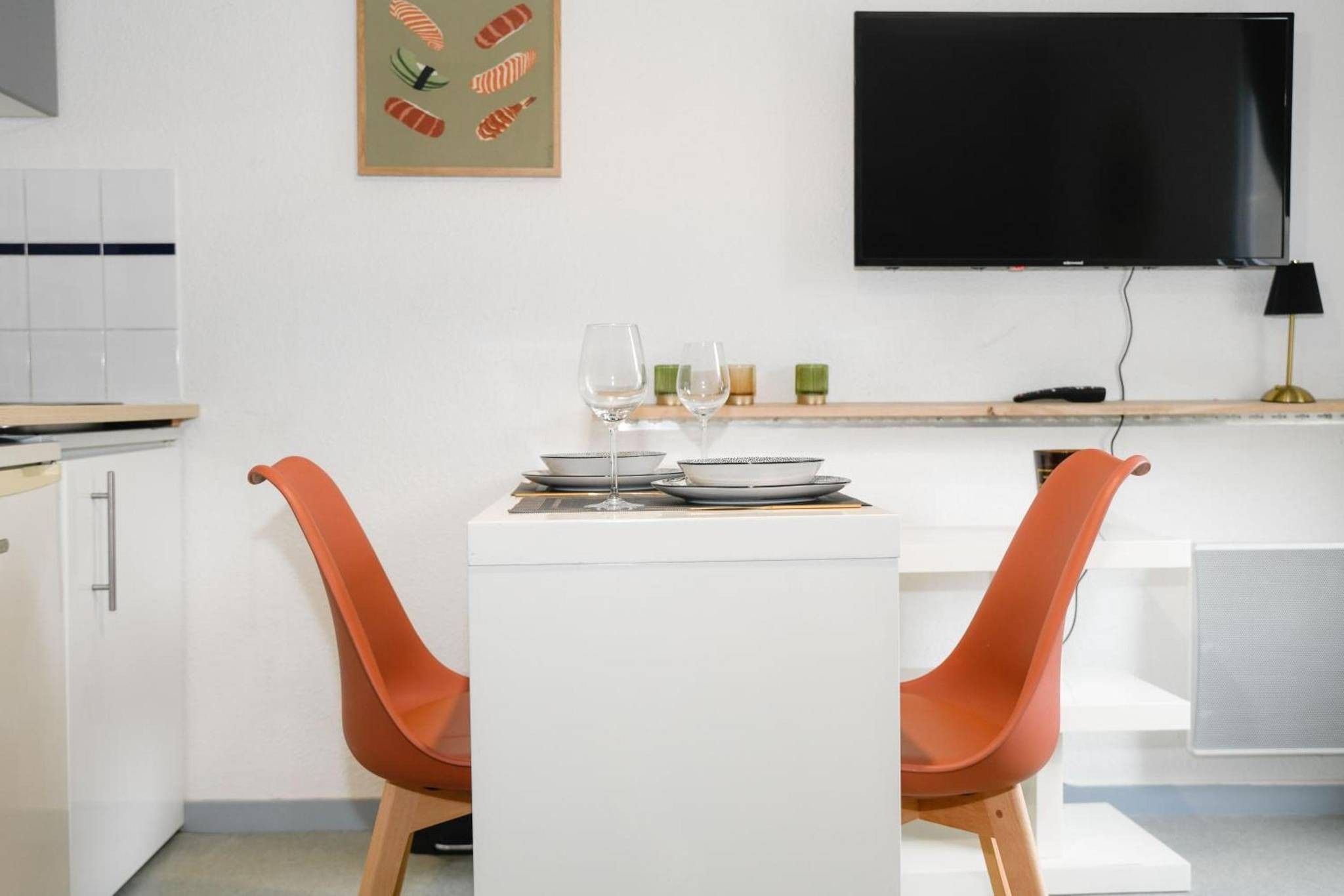 Una cucina moderna con mobili bianchi, una TV nera montata a parete e un tavolo da pranzo con due sedie arancioni.