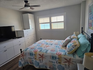 Un dormitorio con cama, televisor, ventilador de techo y ventana con persianas.