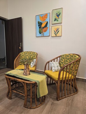 Une pièce comprenant deux chaises en bois avec des coussins jaunes, une petite table recouverte d’une nappe verte et trois œuvres d’art encadrées accrochées au mur.