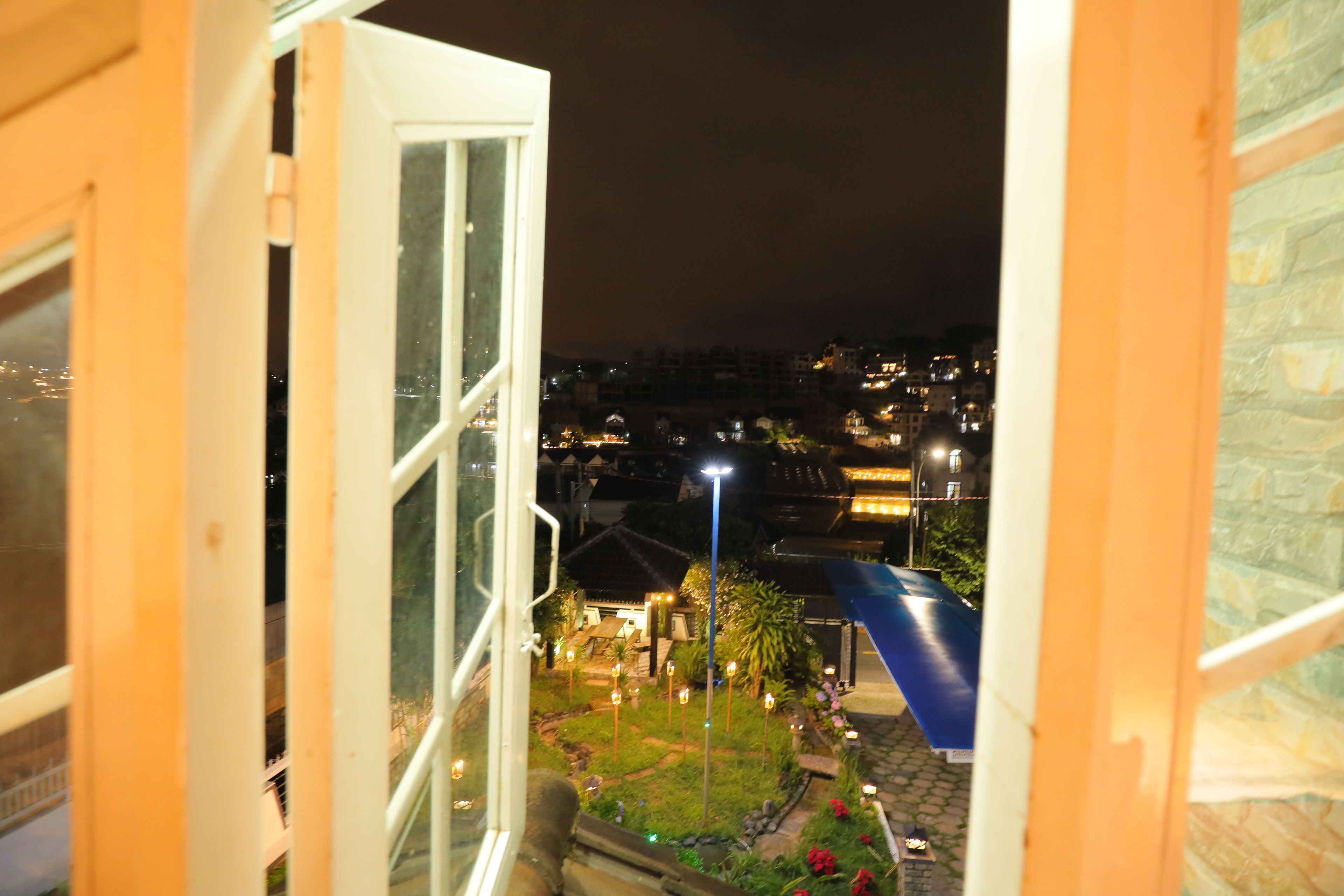 Ein offenes Fenster mit Blick auf einen Innenhof bei Nacht, mit einer blauen Rutsche, Straßenlaternen und Gebäuden in der Ferne.