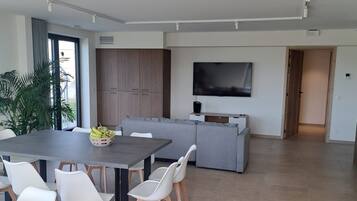Een moderne woonruimte met een eethoek, een grijze bank, een flatscreen televisie en een groot raam.