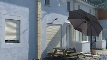 Een gebouw met een witte gevel, een houten picknicktafel en een grote parasol.