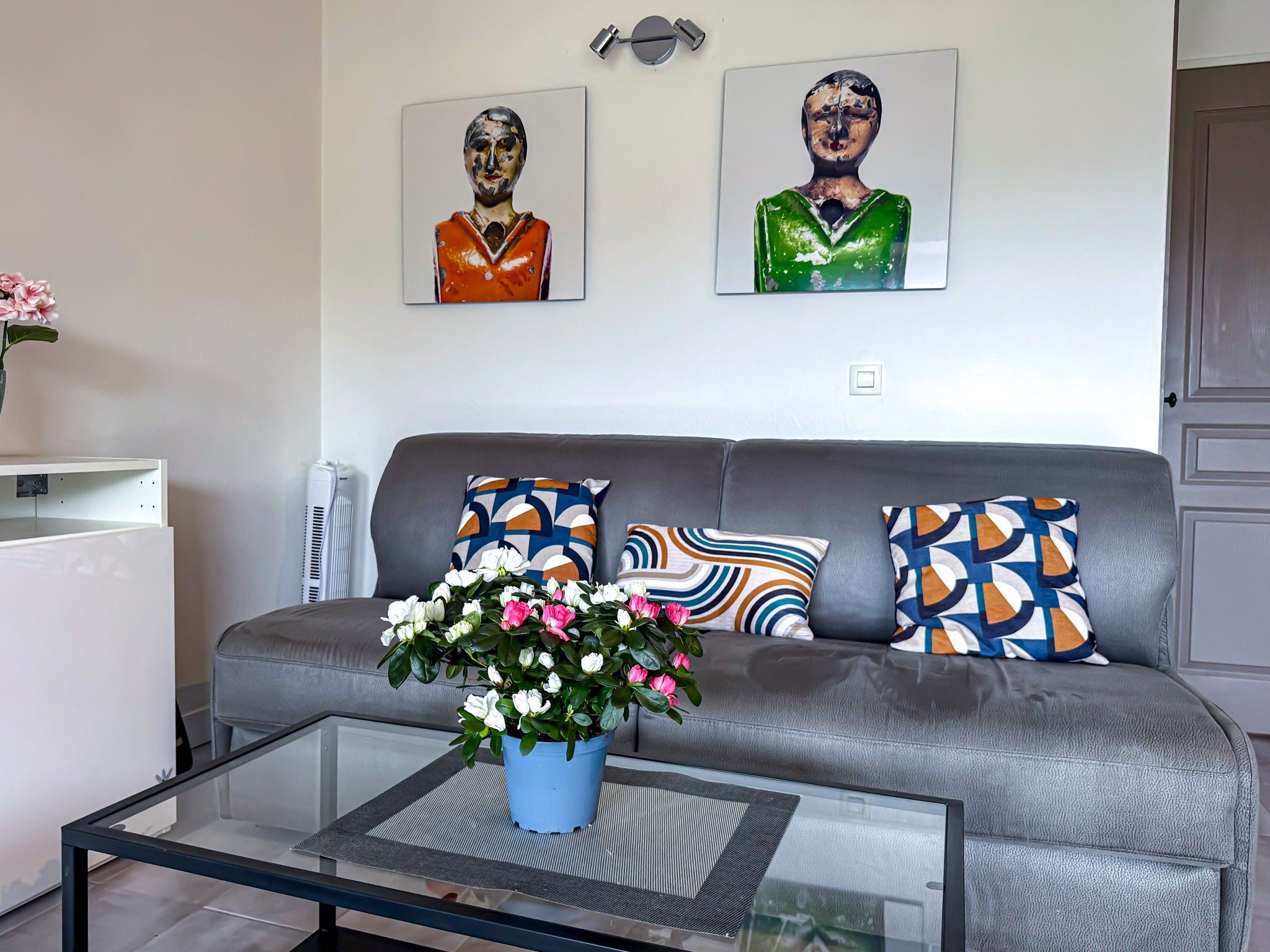 A modern living room with a grey sofa, colorful cushions, a glass coffee table with a flower vase, and two abstract paintings on the wall.