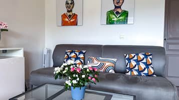 A modern living room with a grey sofa, colorful cushions, a glass coffee table with a flower vase, and two abstract paintings on the wall.