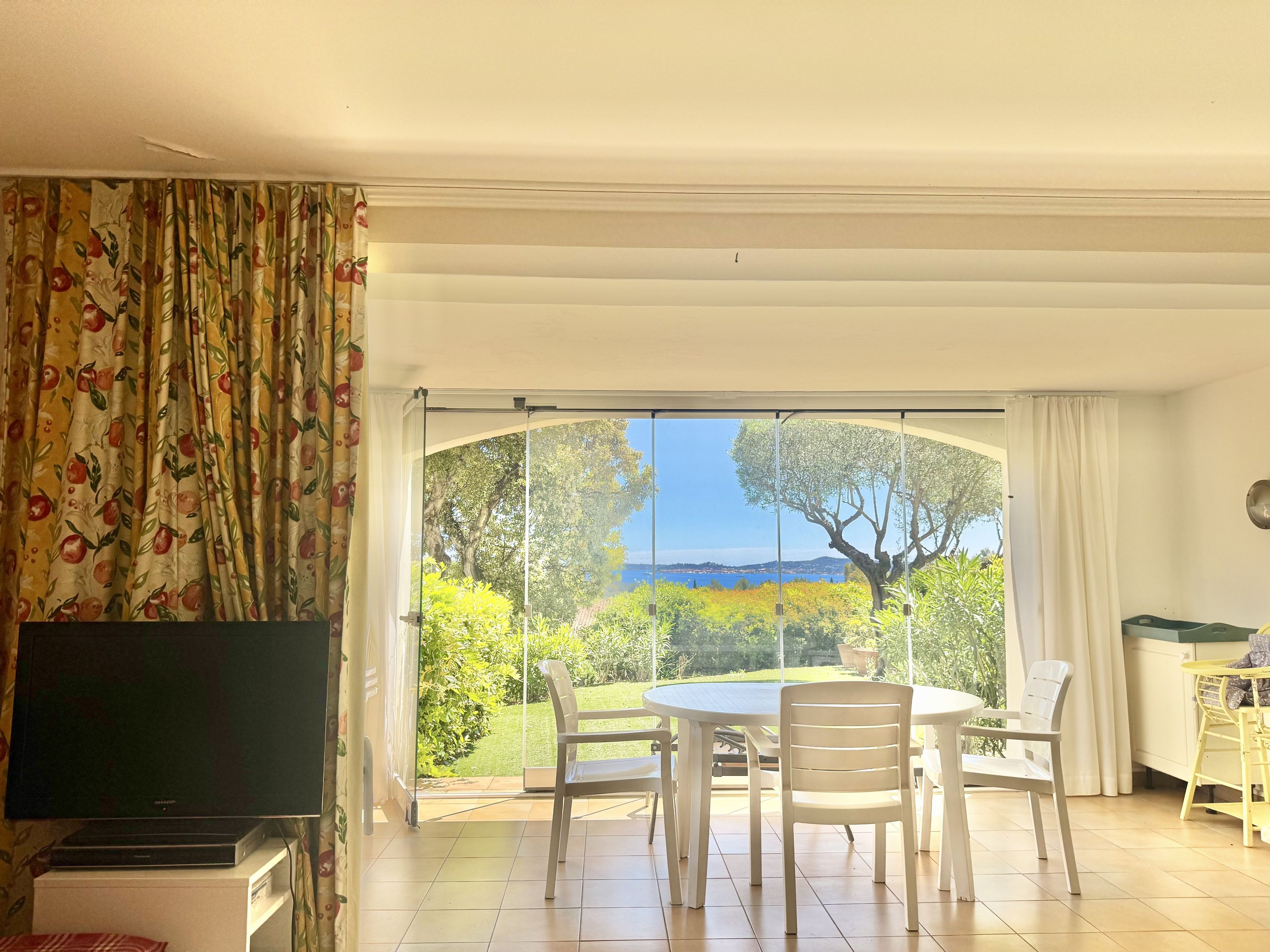 Une pièce dotée d’une télévision, d’une table à manger avec des chaises, et offrant une vue sur un jardin et la mer grâce à de grandes portes vitrées.