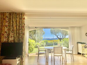 A room with a television, a dining table with chairs, and a view of a garden and sea through large glass doors.