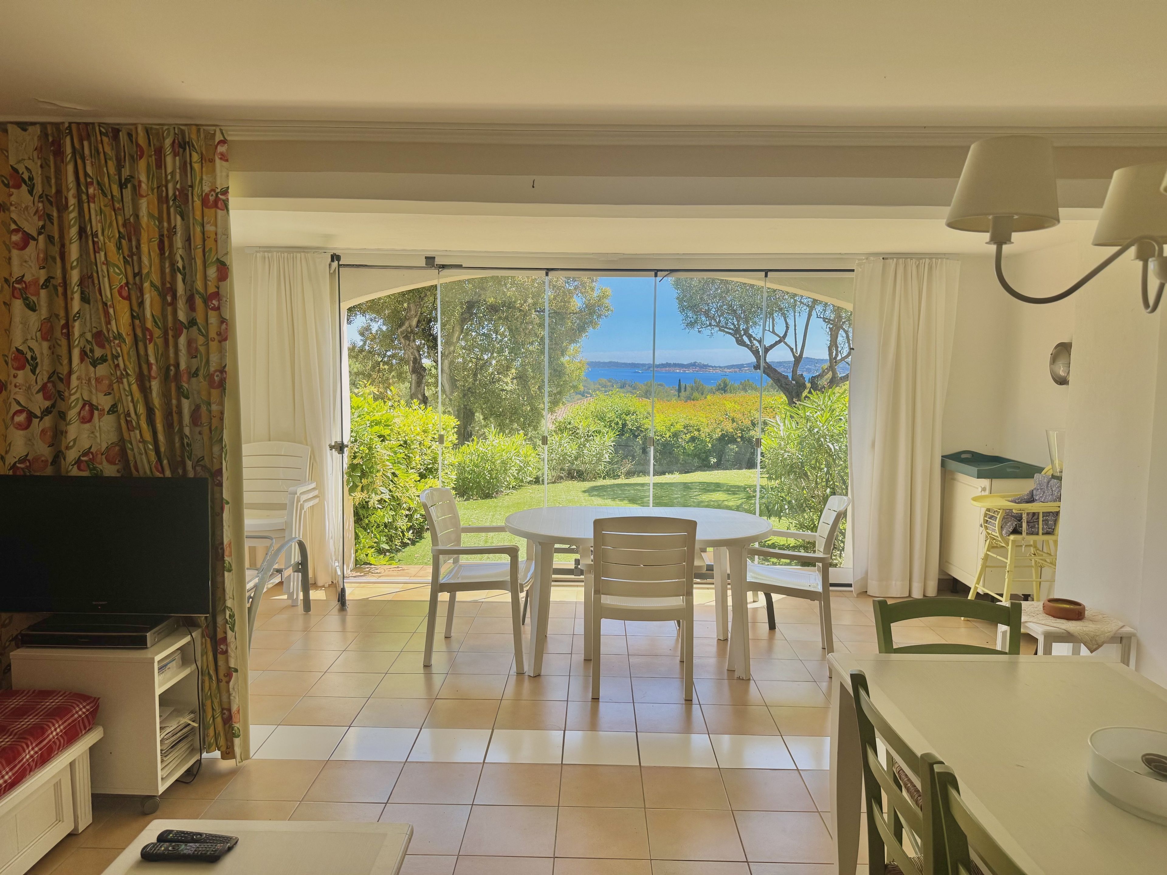 Une pièce comprenant un téléviseur, une table à manger avec des chaises, un petit coin cuisine et une grande porte-fenêtre coulissante donnant sur un jardin avec vue sur la mer.