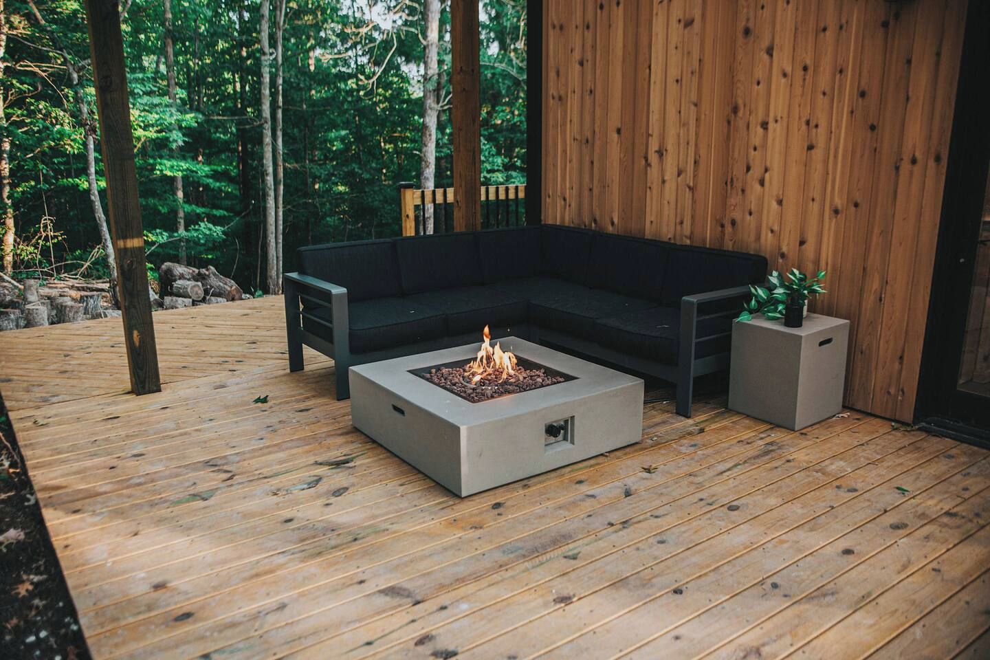 Une terrasse en bois avec un foyer, un canapé noir et un petit pot de fleurs.