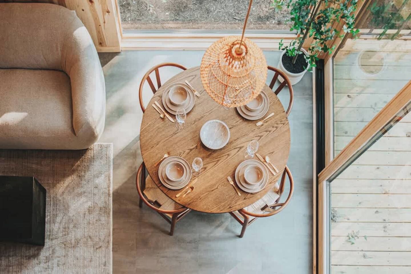 Une table de salle à manger ronde en bois, avec des assiettes, des verres et des couverts, entourée de chaises, dans une pièce dotée d’une grande fenêtre et d’un luminaire suspendu.