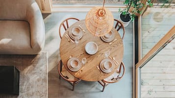 Une table de salle à manger ronde en bois, avec des assiettes, des verres et des couverts, entourée de chaises, dans une pièce dotée d’une grande fenêtre et d’un luminaire suspendu.