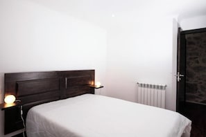 Un dormitori amb un capçal de llit de fusta, un llençol blanc, una taula de nit amb una làmpada i un radiador.