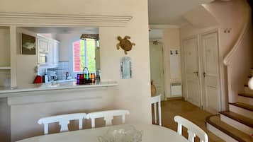A dining area with a white table and chairs, a glass bowl on the table, and a kitchen in the background with white cabinets and a red window.