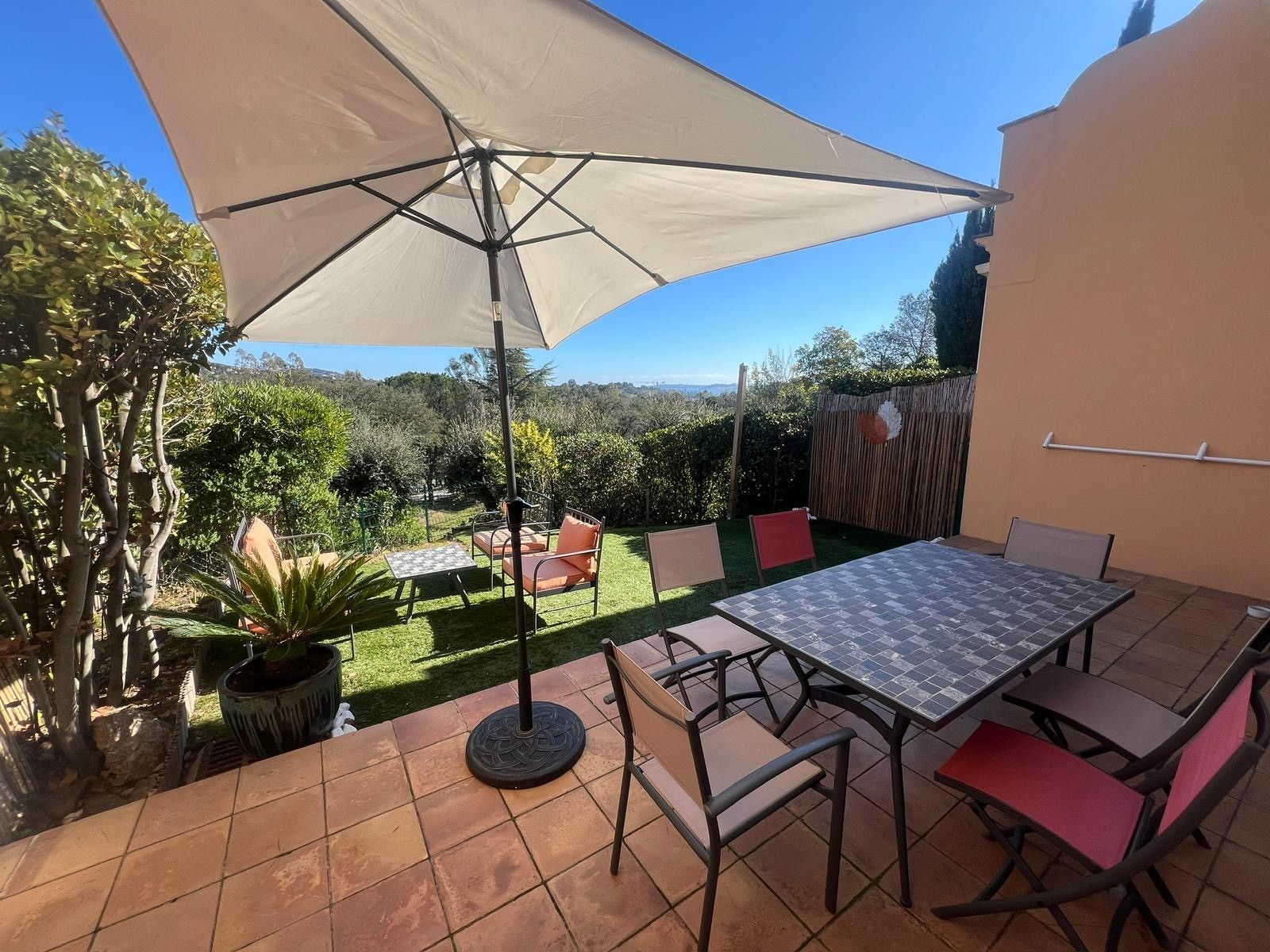 Une terrasse extérieure avec une table et des chaises, un grand parasol et une vue sur le jardin environnant.